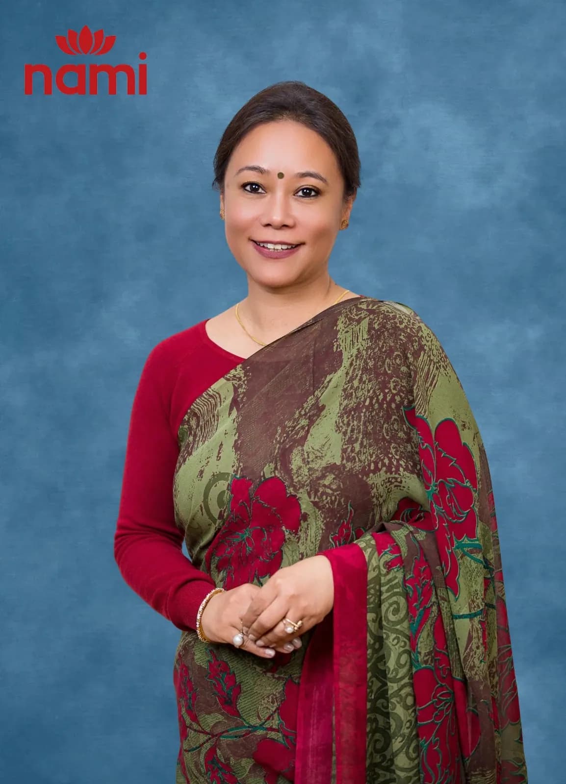 Studio portrait of Ms. Anisha Panday Joshi, Principal of NAMI International School, hands clasped in an olive and red patterned sari over a red blouse against a mottled blue-grey backdrop.