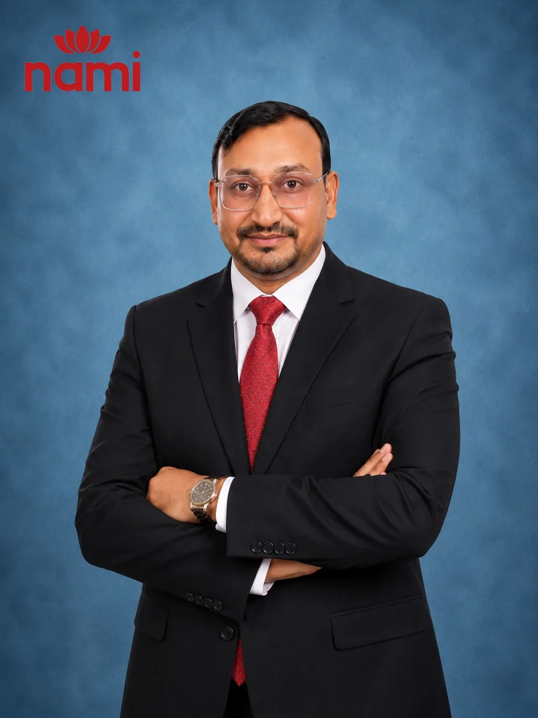 Studio portrait of Mr. Karun Regmi, Head of Business Development and Marketing & Branding at NAMI, arms folded in a black suit, red tie and glasses against a mottled blue-grey backdrop.