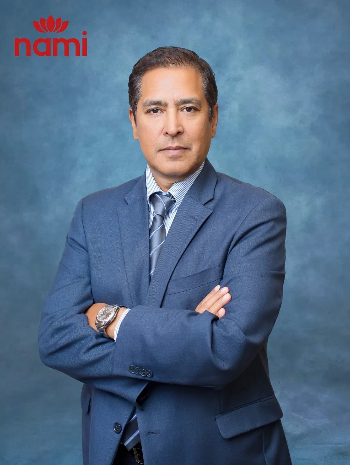 Studio portrait of Mr. Robin Rana, Director at NAMI, arms folded in a slate blue suit and striped grey tie against a mottled blue-grey backdrop.