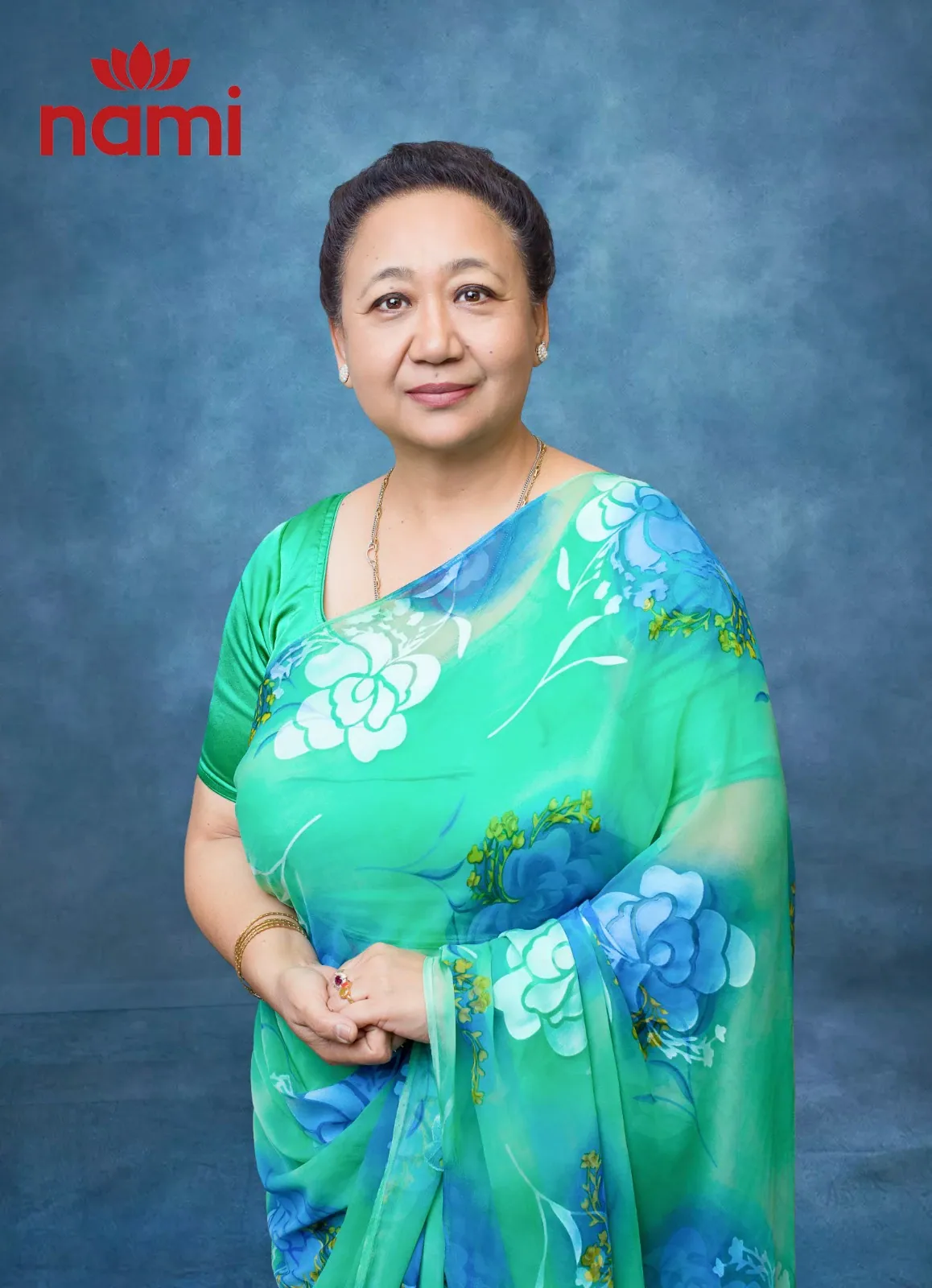 Studio portrait of Ms. Soni Joshi, Director at NAMI, hands clasped in a green and blue floral sari against a mottled blue-grey backdrop.