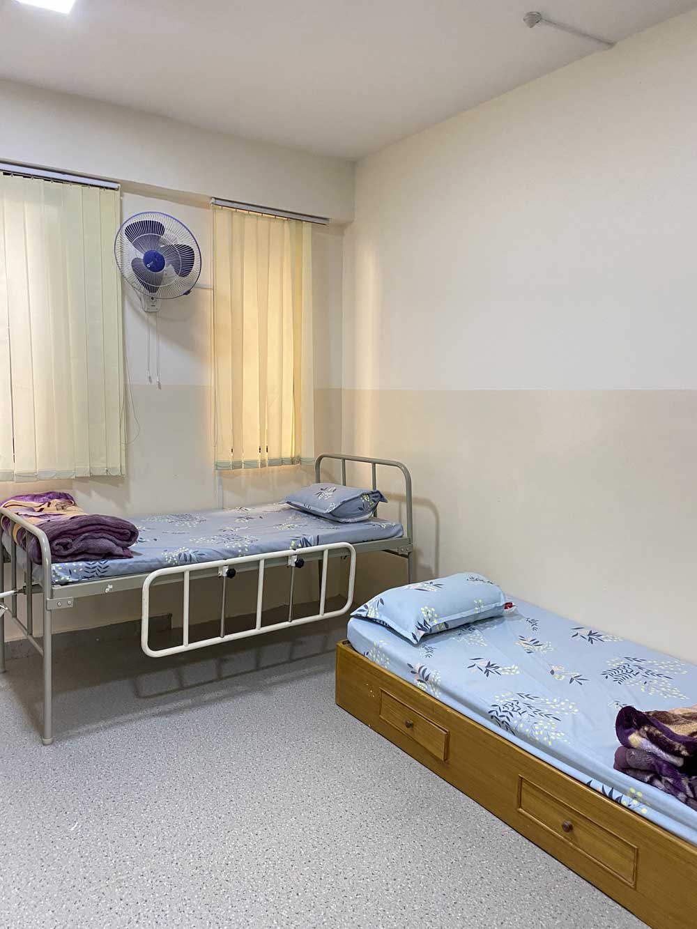 The school infirmary, two single beds made up with pale blue linen and folded blankets beneath a wall fan and a curtained window.