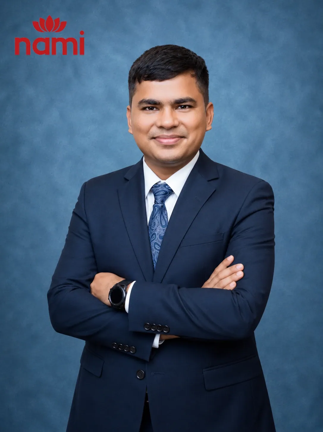 Studio portrait of Mr. Nischal Khadka, Academic Head at NAMI, arms folded in a dark navy suit and patterned blue tie against a mottled blue-grey backdrop.