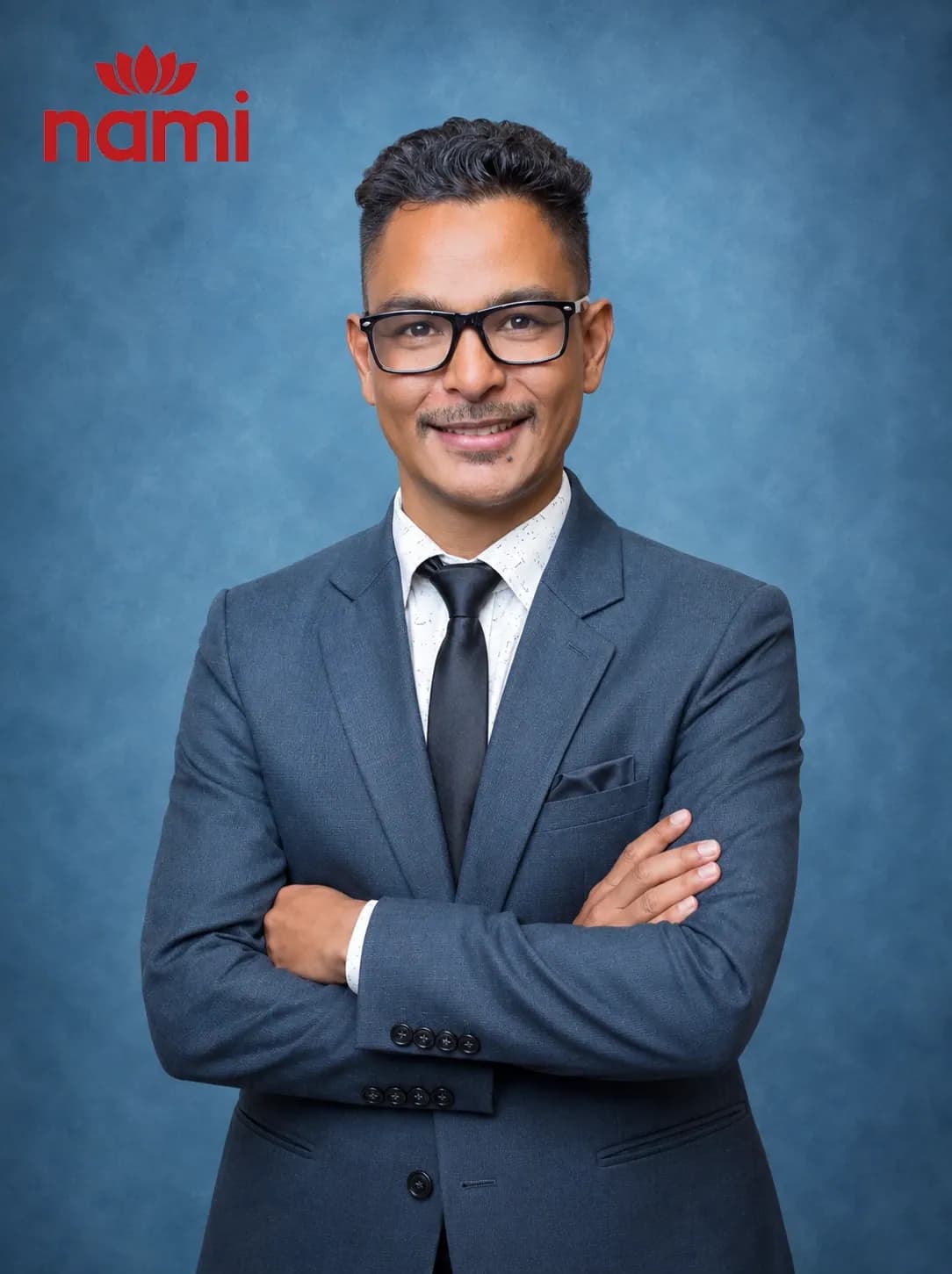 Studio portrait of Mr. Philip Badikar Hilario, A Level Principal at NAMI College, arms folded in a charcoal suit and black tie against a mottled blue-grey backdrop.