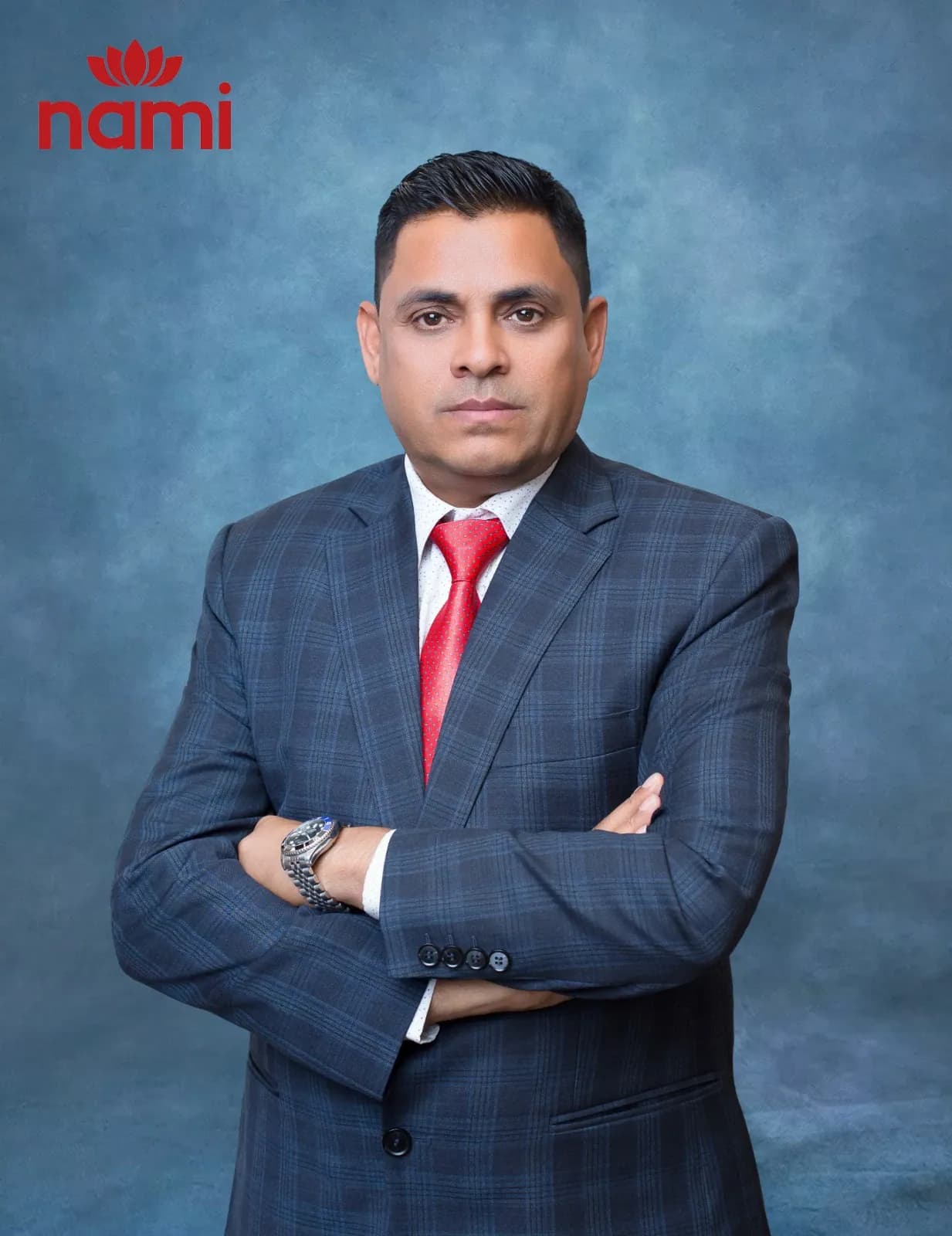 Studio portrait of Mr. Ramesh Prasad Tiwari, Director at NAMI, arms folded in a dark checked suit and red tie against a mottled blue-grey backdrop.