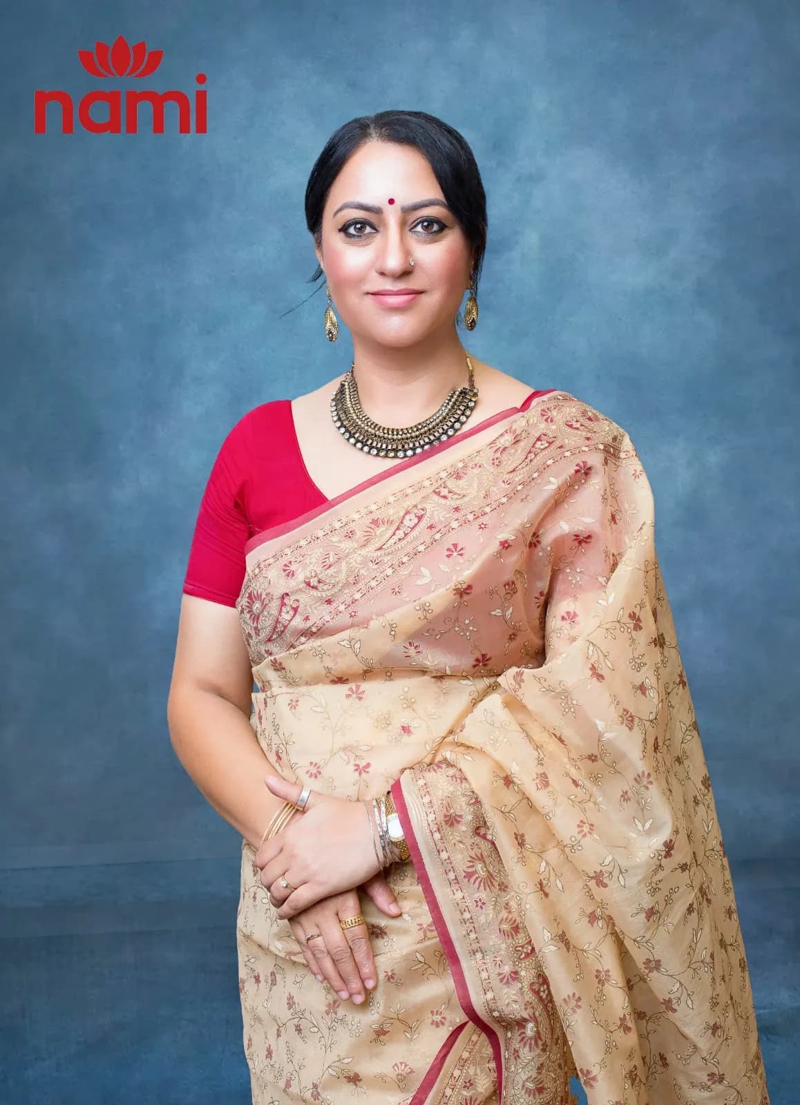 Studio portrait of Ms. Samjhana Phuyal, Director at NAMI, hands clasped in a cream embroidered sari over a red blouse against a mottled blue-grey backdrop.