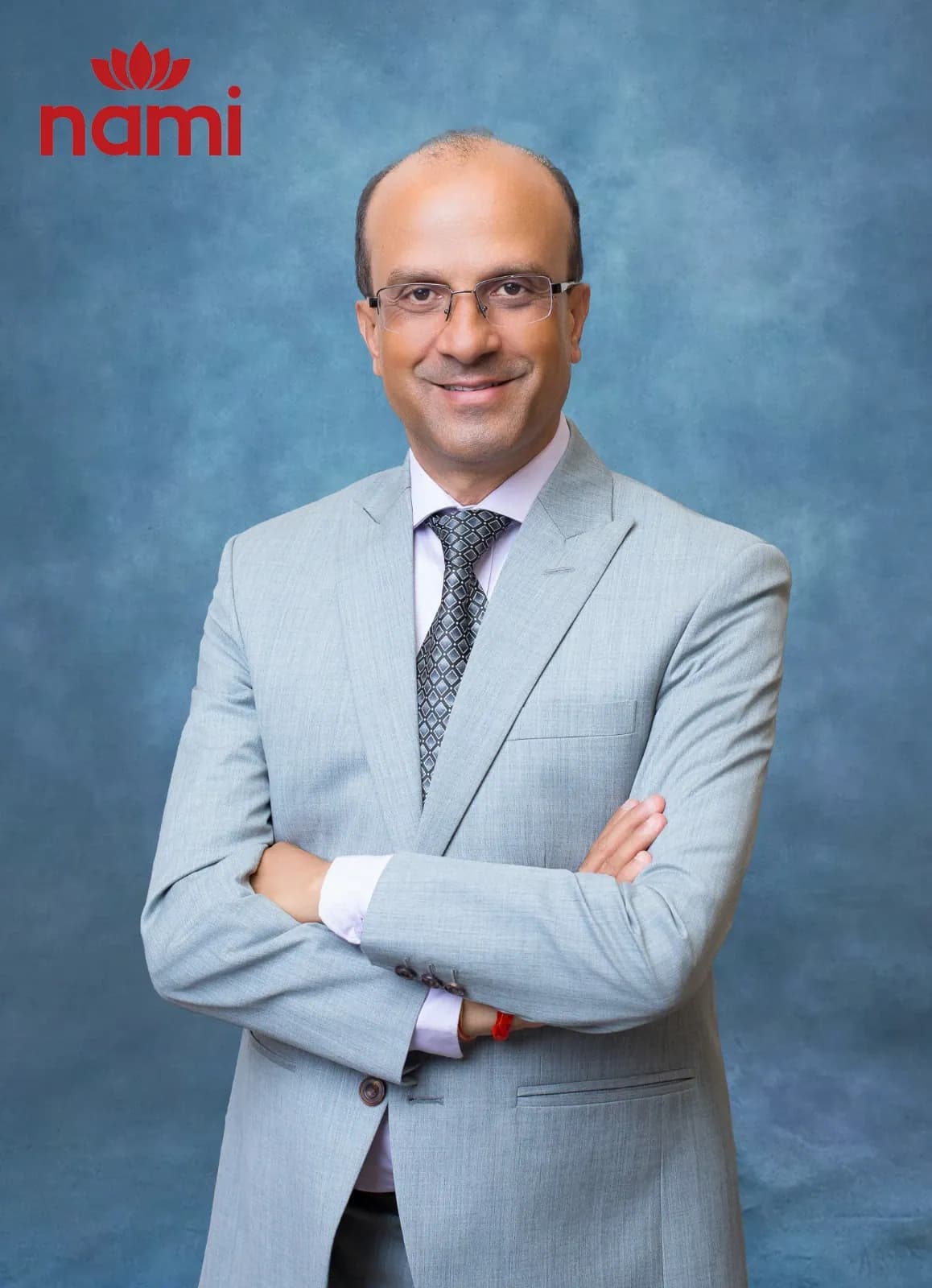Studio portrait of Mr. Suresh Raj Ghimire, Director at NAMI, arms folded in a light grey suit and patterned tie against a mottled blue-grey backdrop.