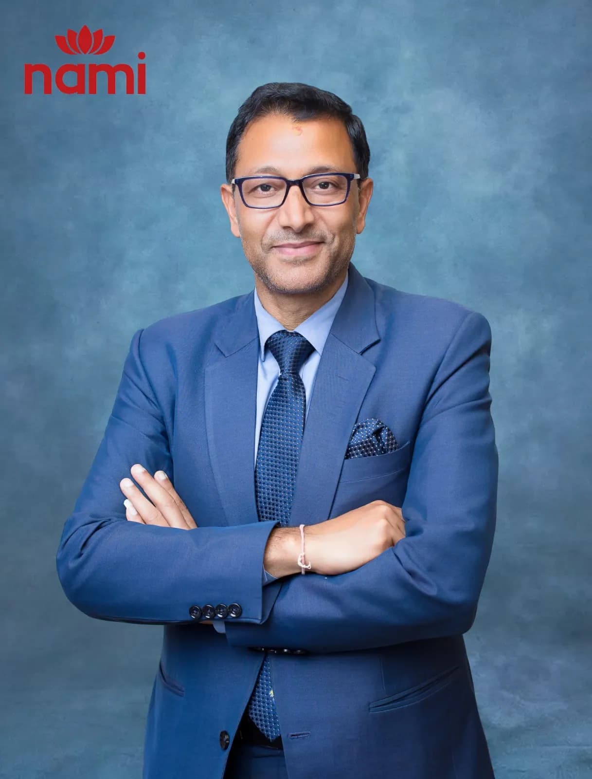 Studio portrait of Mr. Yog Raj Kandel Sharma, Director at NAMI, arms folded in a navy suit and blue tie against a mottled blue-grey backdrop.
