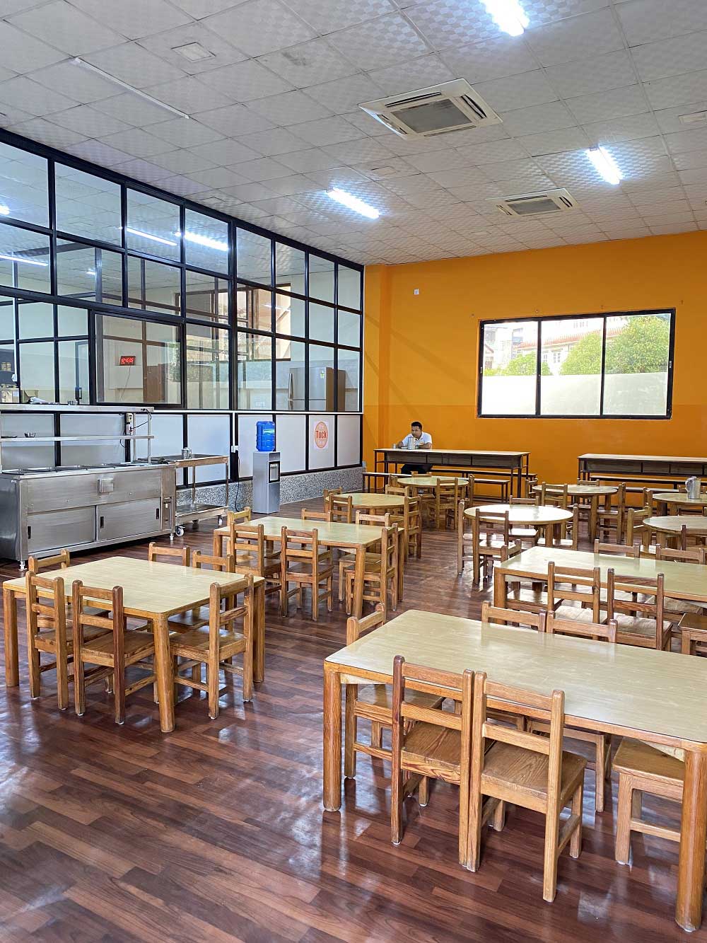 The school cafeteria, wooden tables and chairs ranked across a polished floor beside a glazed partition, with a stainless steel serving counter and a water dispenser against the orange wall.