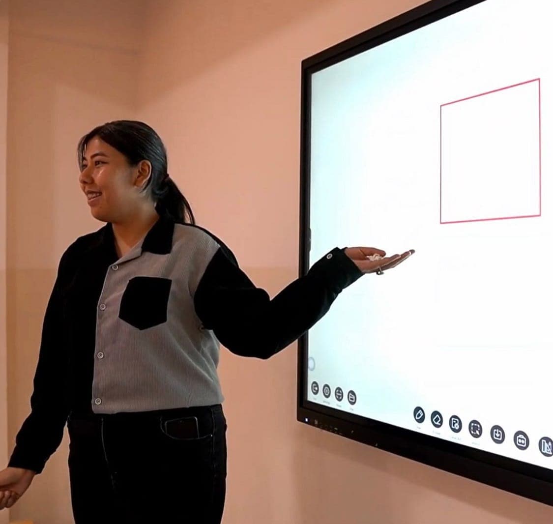 A teacher mid-lesson at a wall-mounted interactive board, presenting a red quadrilateral she has drawn on its touchscreen.