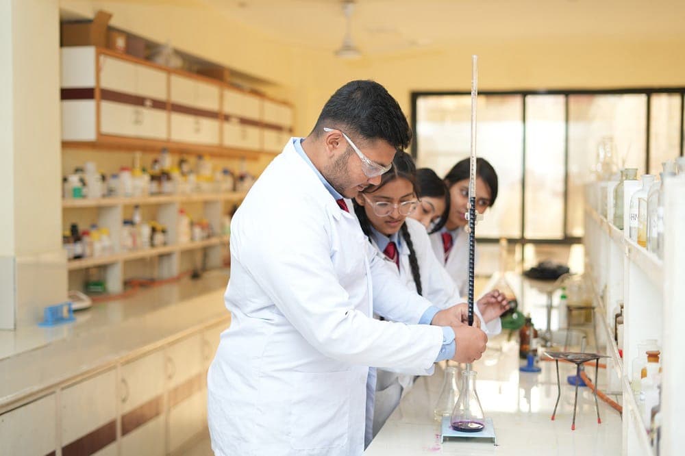 Students in white lab coats and safety goggles running a titration into a conical flask at a laboratory bench, reagent bottles ranked on the shelves behind them.